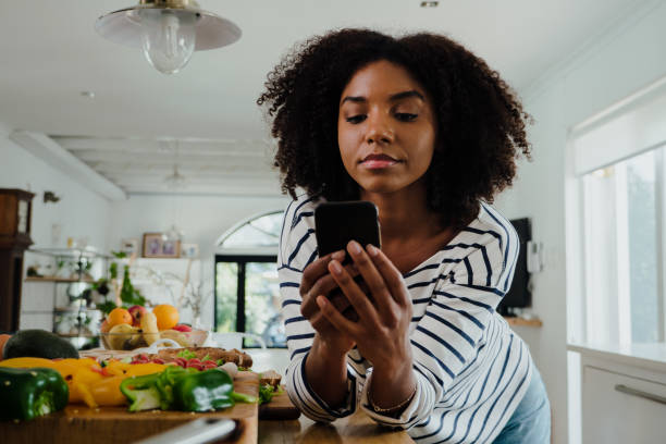 Beautiful focussed African American female researching lunch recipe on smartphone standing next to chopped vegetables in organised kitchen. High quality photo